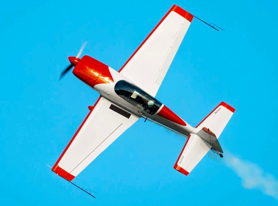 Red and white Extra 300 aerobatic aircraft banking sharply against a bright blue sky, trailing smoke.