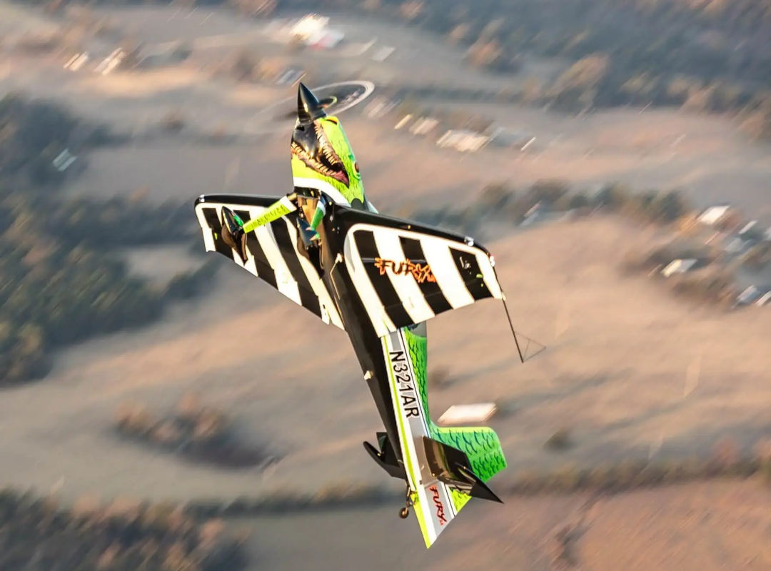 Green, black, and white GB1 GameBird aerobatic aircraft performing a steep, nose-down maneuver over the landscape below.