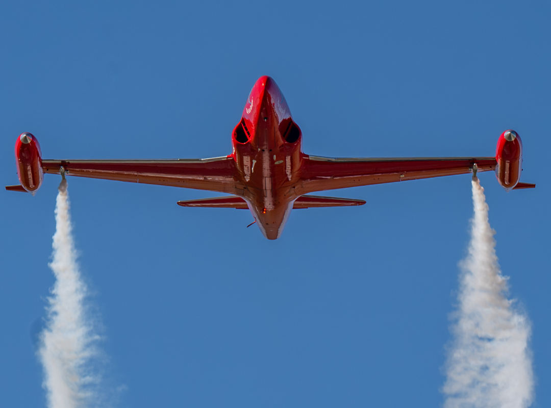Bright red T-33 jet viewed from below in flight, releasing twin white smoke trails against a clear blue sky.